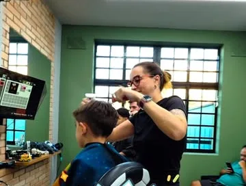 Sala infantil de parede verde: barbeira corta o cabelo de um menino, com um videogame na tela ao lado